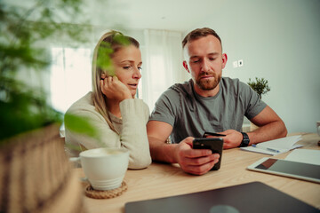 Caucasian couple sitting at home at kitchen table typing on cellular device
