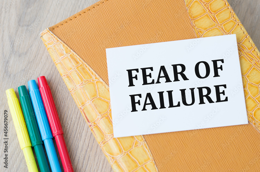 Aufkleber text fear of failure on a white card that lies on a yellow notepad on the table.