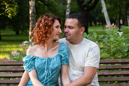 Loving couple on a park bench look at each other with tenderness. Man and a curly woman in a blue dress on a date. Family summer walk.