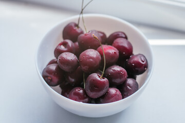Partially blurred white bowl with ripe red sweet cherries, on white surface