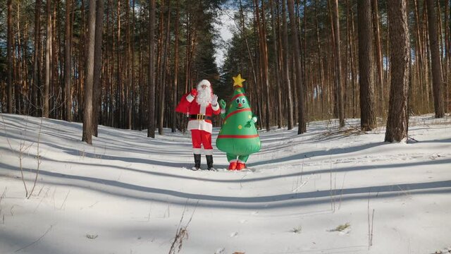 Santa Claus with a funny doll inflatable Christmas tree happily waving his hands looking at the camera while standing in the middle of a snowy winter sunny forest