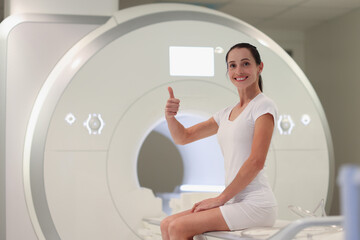 Young woman sitting on magnetic resonance imaging machine and showing thumb up