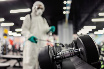 Experts warn of the importance of disinfecting high-risk places in an indoor gym. Man in protective clothing cleans dumbbells and training equipment