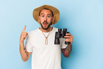 Young caucasian man with tattoos holding binoculars isolated on blue background  having some great idea, concept of creativity.