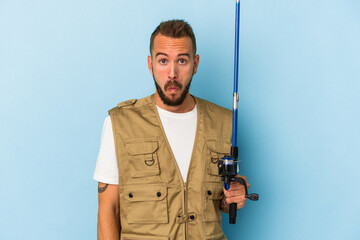 Young caucasian fisherman with tattoos holding rod isolated on blue background  shrugs shoulders and open eyes confused.