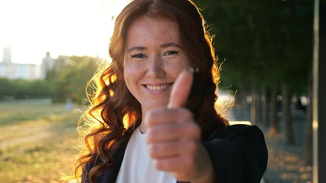 Happy young woman with long loose curly hair and freckles shows thumb-up at bright backlit on city park road with meadow closeup