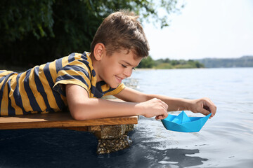 Cute little boy playing with paper boat on wooden pier near river