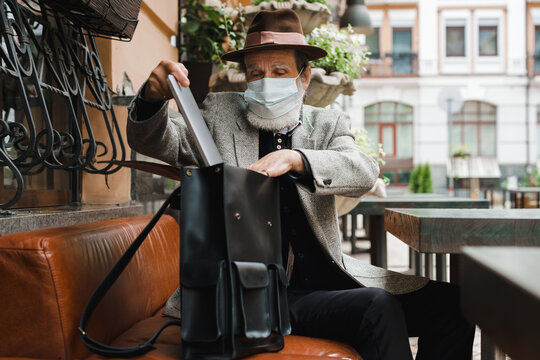 White senior man in face mask getting out laptop of his bag at cafe