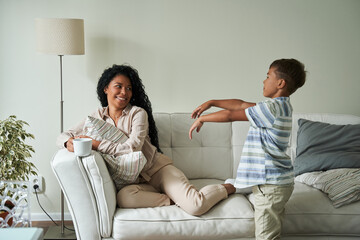 Little boy joking and playing with his mother sitting at the sofa with cup