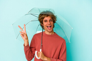 Young caucasian man with makeup holding umbrella isolated on blue background  joyful and carefree...