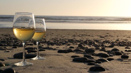 Two wineglasses on sandy ocean beach. Couple of glasses and pebbles, white wine for romantic date near sea water. Cozy beachfront sunset near tide waves. Seamless looped cinemagraph. California, USA.
