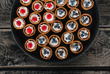Many round dough tartlets with cream, cherries, blueberries are on a tray, plate on a wooden table.