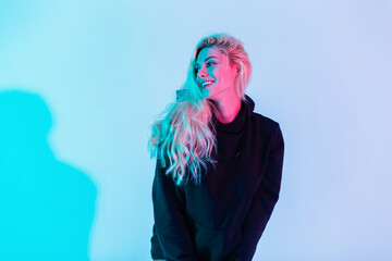 Creative happy portrait of smiling beautiful blonde woman in fashion clothes with black hoodie in studio on multicolored pink and blue background