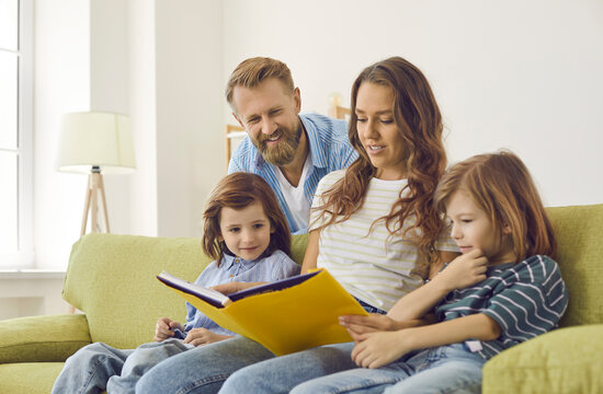 Happy family reading book together. Mom, dad and two little children sitting on soft comfy green sofa at home, looking through photo album, encyclopedia or book of interesting stories and fun facts