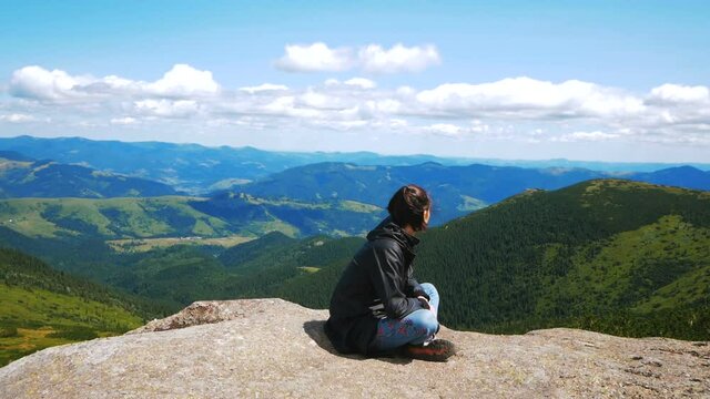 A young woman traveler has reached the top of the mountain and enjoys a beautiful view of the forest and mountains. Young hiker woman smiling and looking at the camera sitting on top of a cliff on the