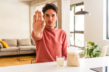 Young mixed race man eating oatmeal and milk for breakfast in his kitchen standing with outstretched hand showing stop sign, preventing you.