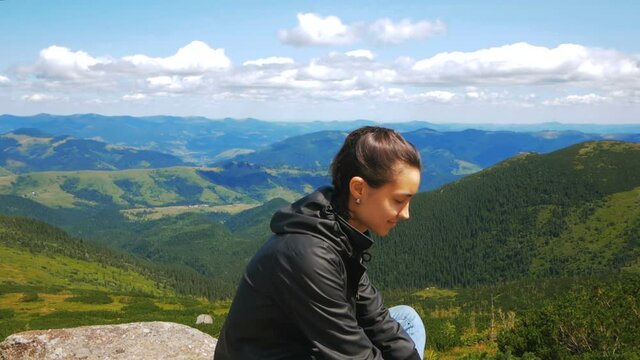 A young woman traveler has reached the top of the mountain and enjoys a beautiful view of the forest and mountains. Young hiker woman smiling and looking at the camera sitting on top of a cliff on the