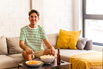 Fototapeta na wymiar Young mixed race man eating popcorns sitting on the sofa happy, smiling and cheerful.