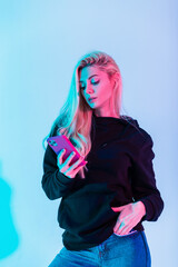 Fashion beautiful young woman in fashionable black hoodie with jeans uses smartphone in multicolored neon pink light in studio