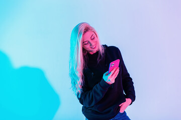 Happy young woman with smile in black hoodie and blue jeans with smartphone typing message in studio on neon pink light background