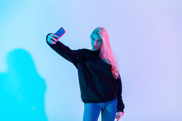 Beautiful woman in trendy casual clothes hoodie and jeans makes selfie on the phone in the studio...