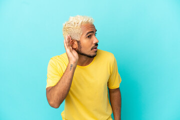 Young Colombian handsome man isolated on blue background listening to something by putting hand on the ear
