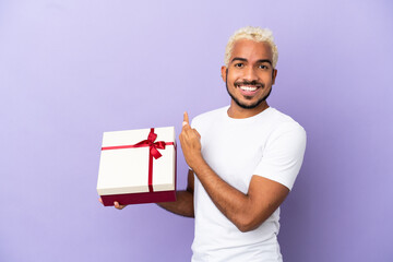 Young Colombian man holding a gift isolated on purple background pointing back