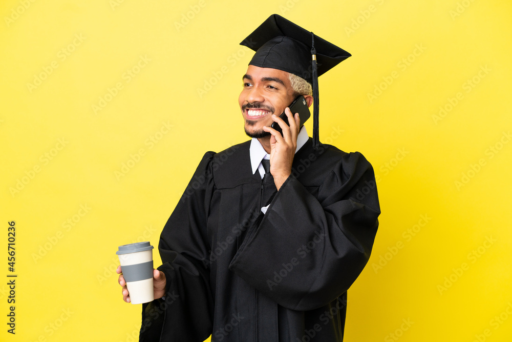 Wall mural Young university graduate Colombian man isolated on yellow background holding coffee to take away and a mobile