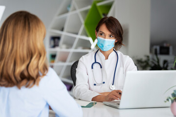 Female doctor wearing face mask and consulting her patient while sitting at desk in doctor's office