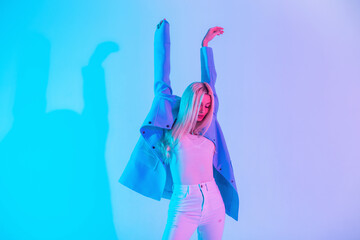 Young stylish beautiful dancer woman in fashionable elegant outfit with a blue blazer, white blouse and jeans is dancing in pastel colored neon pink lights