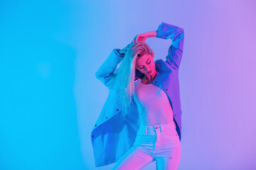 Glamorous stylish young blonde woman model in fashionable suit with blue blazer with white jeans poses on bright pastel colored neon pink lights in studio