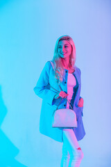 Happy blonde model girl with a white smile wearing fashionable elegant clothes with a blue blazer, white tank top and jeans holding a white bag in the studio on a pastel colored pink neon light