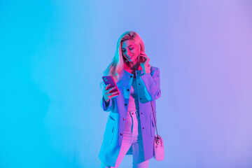 Beautiful young happy woman in a bright fashionable business suit look with a stylish handbag holds a phone and uses a mobile application in the studio on a colored neon pink light