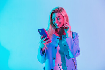 Happy fashionable young girl with a beauty smile in fashion elegant business clothes with a jacket holding a smart phone and typing a message on a multicolored neon pink light in the studio - obrazy, fototapety, plakaty