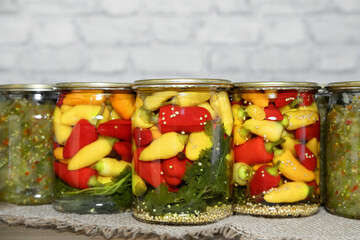 Mini chili peppers of red, yellow and orange colors, canned in glass jars with herbs, on a wooden table. Homemade pickled peppers in Asian style. Preparations for the winter. Copy space