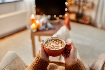 people and leisure concept - young woman watching tv and drinking hot chocolate with marshmallow with her feet on table at cozy home