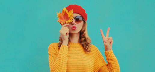 Autumn portrait of beautiful young woman with yellow maple leaves blowing her lips wearing a knitted sweater, red beret on blue background