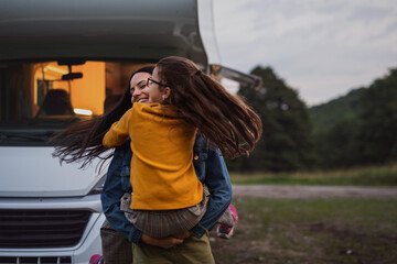 Happy mother with small daughter hugging by car outdoors in campsite at dusk, caravan family...