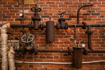 Old rusty water pipes with shut-off valves and sensors in an industrial building opposite a brick wall.