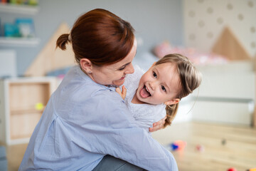 Happy mother with small daughter playing indoors in bedroom, laughing.