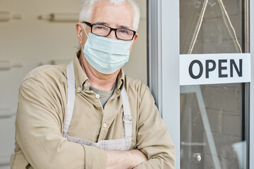 Portrait of mature Caucasian owner of store or cafe in mask leaning on doorway, open signboard on door