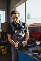 At car service station. One young man, male auto mechanic in dungarees working at car service station, indoors. Concept of labor, occupation, business, caree, job