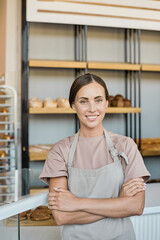 Portrait of happy young Caucasian owner of bakery standing with crossed arms against display with loaves of bread