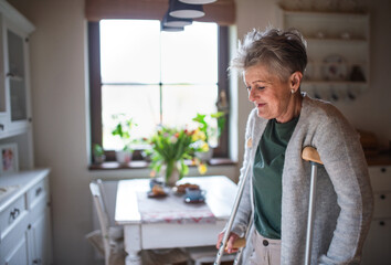 Senior woman with crutches indoors at home, walking in kitchen.