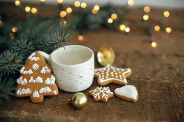 Christmas gingerbread cookies, coffee cup, fir branches and bauble on background of warm lights bokeh on rustic wooden table. Moody atmospheric image. Winter countryside hygge. Seasons greeting