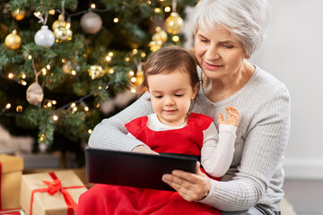 winter holidays and family concept - happy grandmother and baby granddaughter with tablet pc computer sitting near christmas tree at home