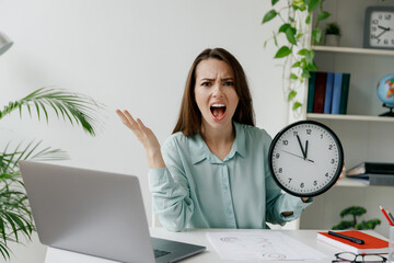 Young angry stressed employee business woman in blue shirt hold in hands clock spread arms scream sit work at workplace white desk with laptop pc computer at office indoor Achievement career concept.