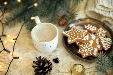 Obraz na płótnie Canvas Homemade christmas gingerbread cookies, warm coffee in stylish cup, fir branches and warm lights on cozy knitted background. Atmospheric festive time and hygge home. Happy Holidays. Hello winter