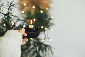 Hand in cozy sweater holding vintage candlestick with burning candle on background of warm lights,...