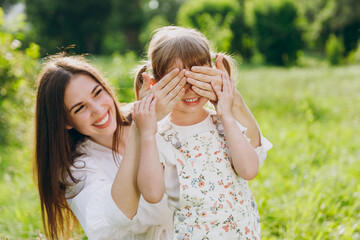 Obraz premium Smiling young woman in white clothes close eyes with hands play guess who or hide and seek with child baby girl 5-6 years old Mommy rest with little kid daughter outdoor together. Love family concept